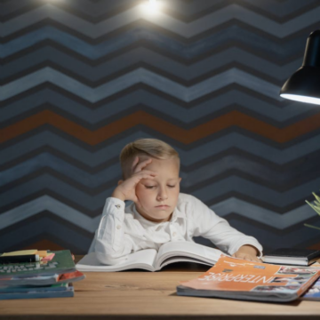 A kid wearing a white shirt is sitting at a desk while reading a book