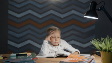 A kid wearing a white shirt is sitting at a desk while reading a book