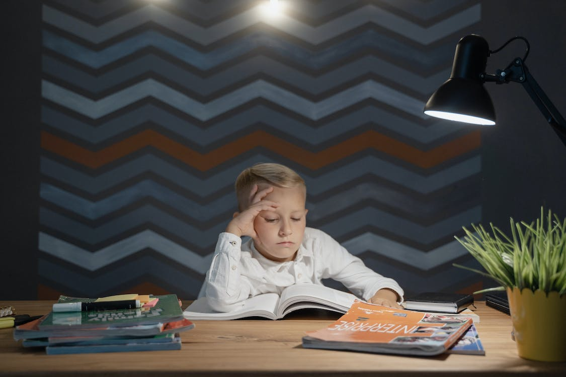 A kid wearing a white shirt is sitting at a desk while reading a book