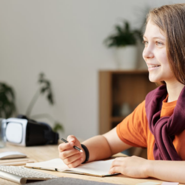 A girl wearing an orange shirt sitting at a computer desk while smiling at the PC screen