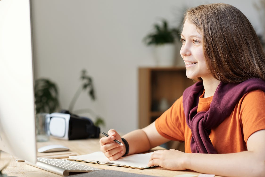 A girl wearing an orange shirt sitting at a computer desk while smiling at the PC screen