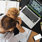 A woman is sitting in front of a MacBook with a notebook lying open as she holds her head in frustration