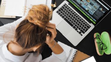 A woman is sitting in front of a MacBook with a notebook lying open as she holds her head in frustration