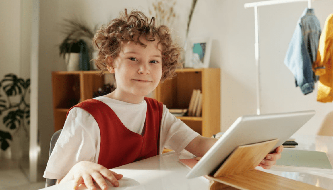 A child sitting down at a desk is holding a silver tablet and smiling while looking at the camera