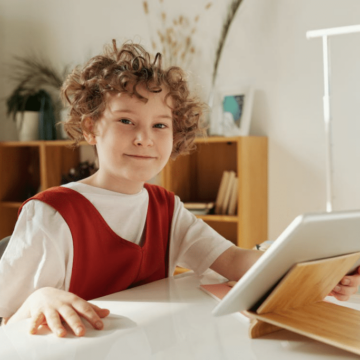 A child sitting down at a desk is holding a silver tablet and smiling while looking at the camera