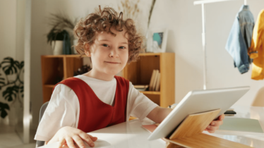 A child sitting down at a desk is holding a silver tablet and smiling while looking at the camera