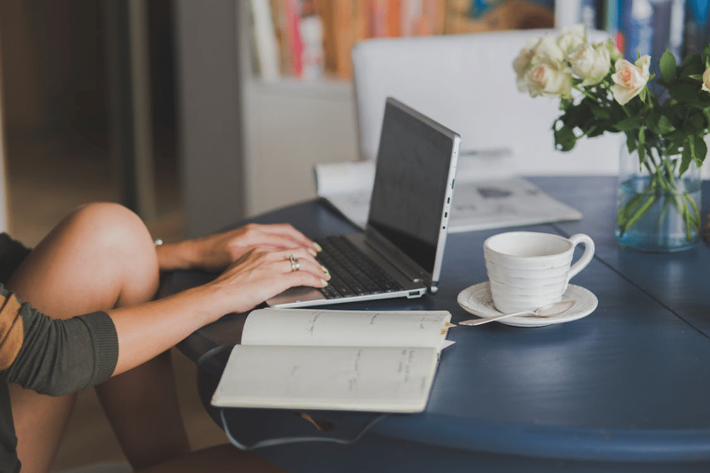 A person sits on a table next to an open notebook and a white tea cup while they work on a silver laptop