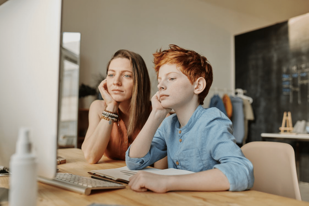 A woman leaning next to a boy who’s sat in front of a PC screen as both look ahead